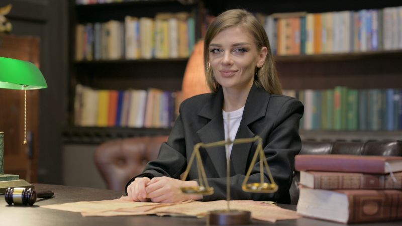A law school graduate in a black blazer sits at a desk with legal books, a gavel, and a scale of justice in front of her, with bookshelves in the background.