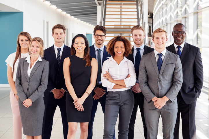 A diverse group of nine professionally dressed lawyers, including minority candidates for legal positions, stand smiling together in a modern office hallway with stairs in the background.