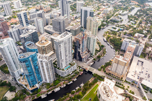 Aerial view of Fort Lauderdale with modern high-rise buildings, a winding river with boats, and lush greenery, showing a mix of urban and natural environments.
