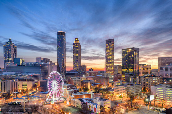 Cityscape of downtown Atlanta at sunset, featuring tall buildings with lights on, a large Ferris wheel in the foreground, and a colorful sky with clouds.