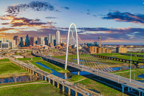 Aerial view of Dallas, Texas at sunset, featuring the Margaret Hunt Hill Bridge in the foreground and the downtown skyline in the background with colorful clouds in the sky.