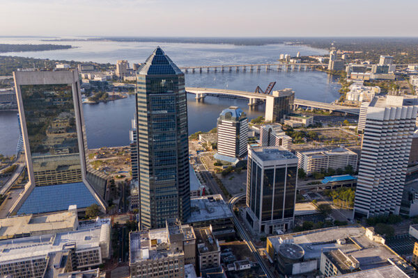 Aerial view of downtown Jacksonville, Florida, with tall modern buildings, the St. Johns River, and bridges stretching across the water under a clear sky.