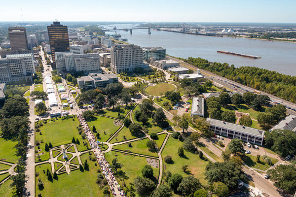 Aerial view of Baton Rouge with office buildings, a river with a barge, a bridge in the distance, and a large park with geometric gardens and walking paths in the foreground.