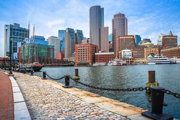 Boston waterfront with cobblestone walkway, docked boats, and a city skyline of modern and historic buildings under a blue sky.