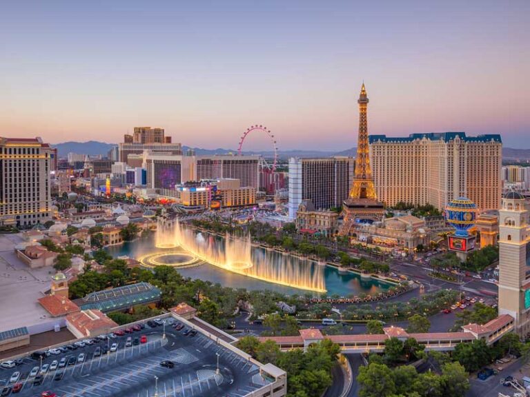 Aerial view of the Las Vegas Strip at sunset, featuring the Bellagio fountains in mid-show, the replica Eiffel Tower, hotels, casinos, and the High Roller Ferris wheel in the background.