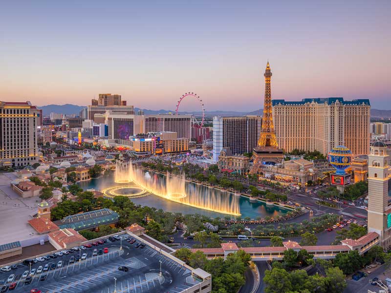 Aerial view of the Las Vegas Strip at sunset, featuring the Bellagio fountains in mid-show, the replica Eiffel Tower, hotels, casinos, and the High Roller Ferris wheel in the background.