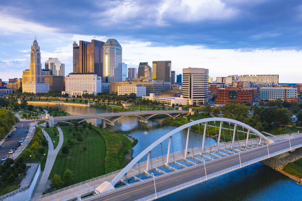Aerial view of downtown Columbus, Ohio, featuring modern skyscrapers, a large arched bridge, and the Scioto River with greenery along the riverbanks under a partly cloudy sky.