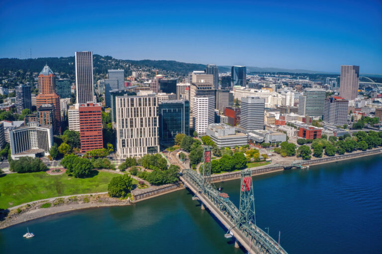 Aerial view of downtown Portland, Oregon, featuring tall buildings, a bridge crossing the Willamette River, and green spaces along the waterfront under a clear blue sky.