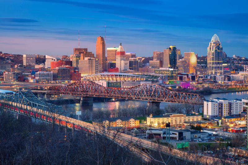 Cincinnati, Ohio skyline at dusk with tall buildings, a lit stadium, and bridges crossing a river; lights reflect on the water, and trees are visible in the foreground.