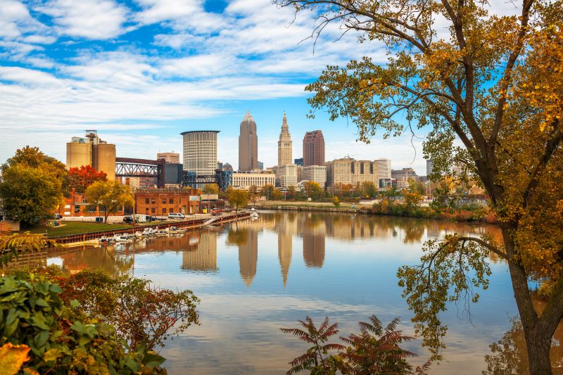 The Cleveland skyline is reflected in the Cuyahoga River, surrounded by autumn trees and a partly cloudy sky. A mix of modern and historic buildings lines the waterfront.