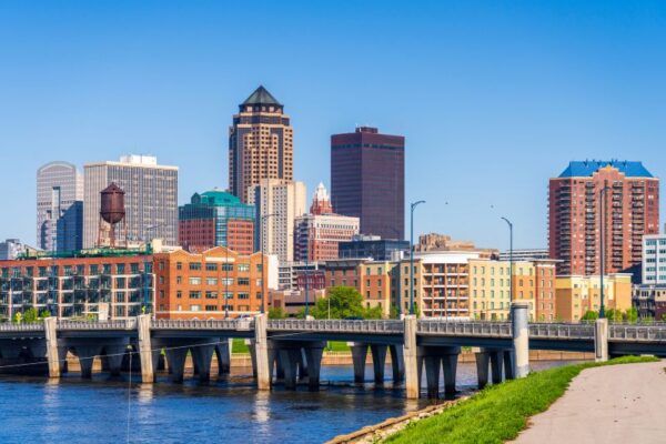 Des Moines Iowa skyline with modern and historic buildings under a clear blue sky, featuring a bridge crossing a river in the foreground and paths lined with greenery along the water.