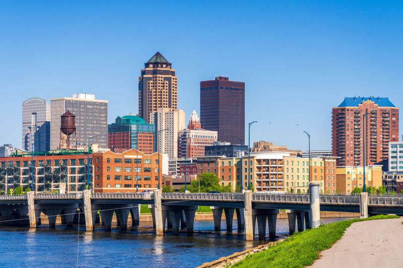 Des Moines Iowa skyline with modern and historic buildings under a clear blue sky, featuring a bridge crossing a river in the foreground and paths lined with greenery along the water.