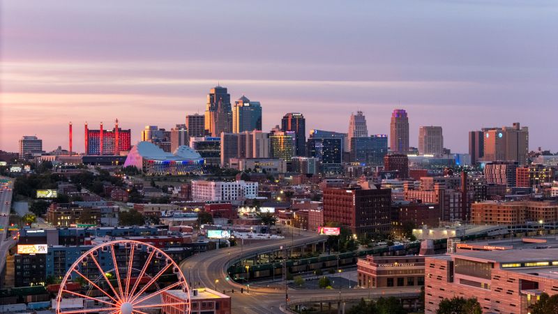 A colorful sunset view of downtown Kansas City Missouri, featuring modern skyscrapers, lit-up buildings, and a ferris wheel in the foreground, with city streets and trees scattered throughout the urban landscape.