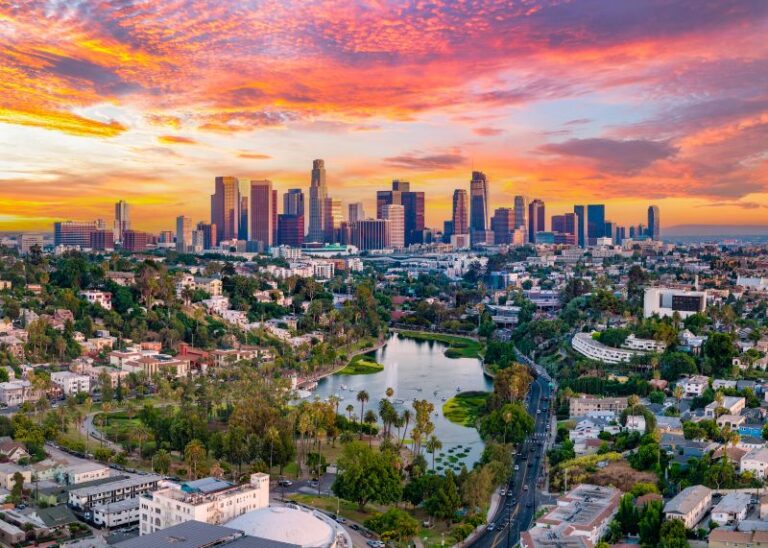 Aerial view of downtown Los Angeles at sunset, with skyscrapers, residential areas, and a park with a lake in the foreground under a vibrant, colorful sky.