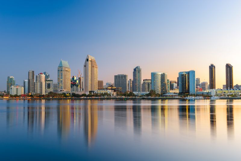 San Diego skyline at sunrise with modern skyscrapers reflected on calm water under a clear blue sky.
