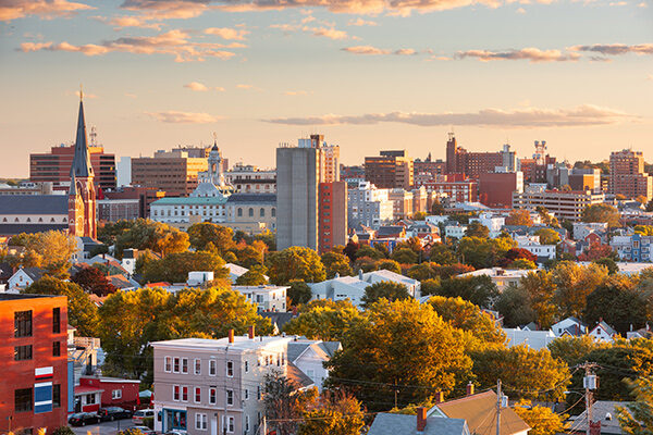 Portland Maine at sunset with a mix of trees, residential houses, and taller downtown buildings under a partly cloudy sky. Warm sunlight highlights autumn foliage and the varied architecture.