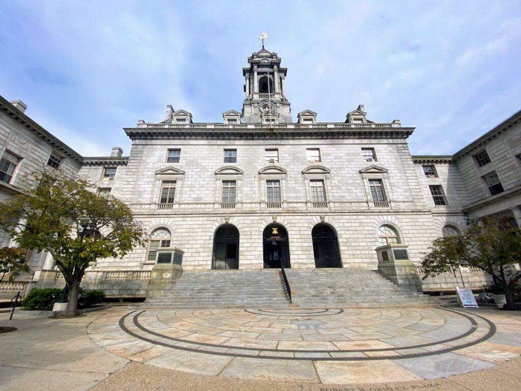 Portland Maine City Hall building with a central clock tower and arched entrance is seen from the front, with a circular stone plaza and stairs leading up to the doors.