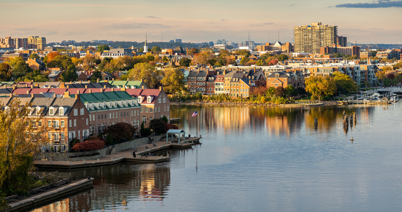 A waterfront neighborhood with brick townhouses and trees lines the edge of a calm river along the Waterfront Of City Of Alexandria, Virginia.