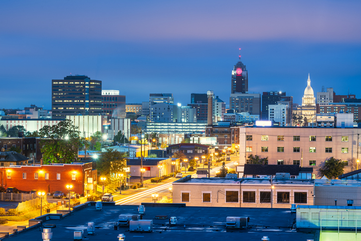 Cityscape of Lansing, Michigan at dusk with buildings lit and the Michigan State Capitol dome visible in the background.