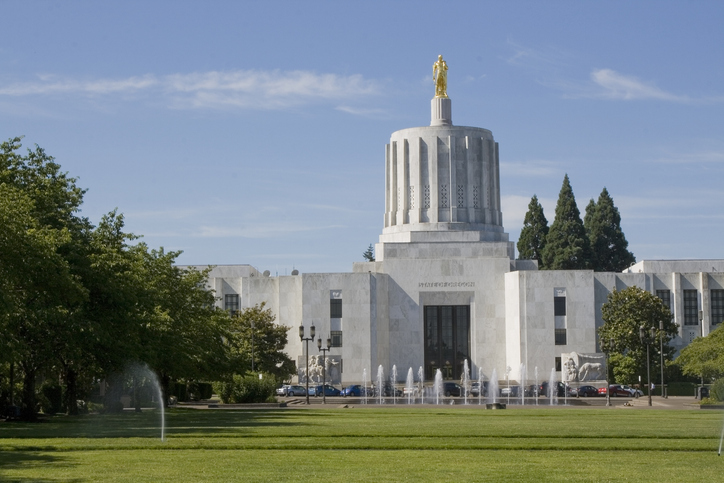 Oregon State Capitol building in Salem, OR with gold statue on top, surrounded by trees, grass, and fountains under a clear sky.