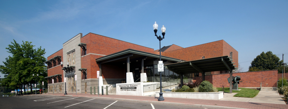 Red brick Deschutes County Courthouse building in Bend, OR with ramp, stairs, and empty parking spaces in front.