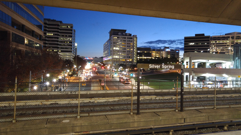 View of downtown Silver Spring at dusk, with buildings, streetlights, cars, and train tracks in the foreground.
